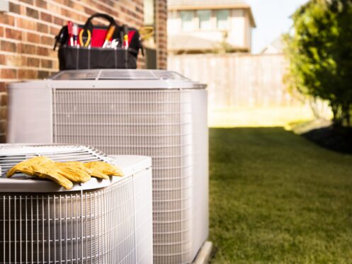 Outdoor residential air conditioning units with work gloves and an HVAC technician’s tool bag ready for AC service.